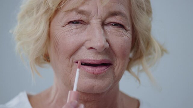 Close-up of beautiful grandmother applying lip gloss on lips preparing for a date. Portrait of smiling older woman doing make-up looking in the mirror on grey background.