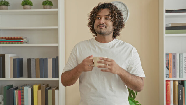 Man holding a coffee mug with both hands, wearing white t-shirt, standing by bookshelf and clock in office; contentment morning break.