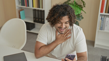 Young indian man holding smartphone, hand to mouth, smiling in a building study with visible bookshelves and laptop  surprise joy. © Krakenimages.com