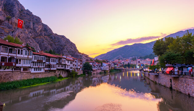 Stunning sunset in Amasya, Turkey. Traditional Ottoman houses along the Yesilirmak River reflecting in the calm water. Majestic mountains and historic city architecture under a vibrant orange sky.