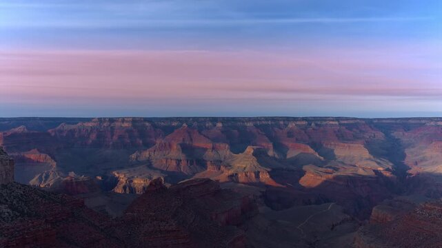 Arizona sunset time-lapse captures the Grand Canyon as golden hour light transitions across ancient layered rock formations. Pink and purple clouds drift over vast canyon depths during twilight.