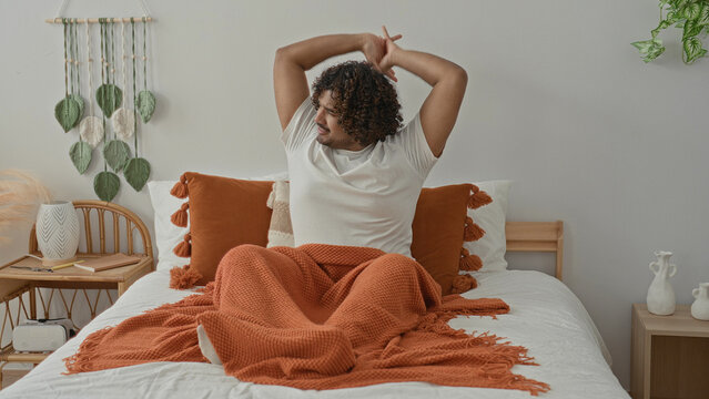 Young indian man on bed stretches arms up, yawning with orange blanket, tassel pillow and wicker bedside table in studio; morning serenity.