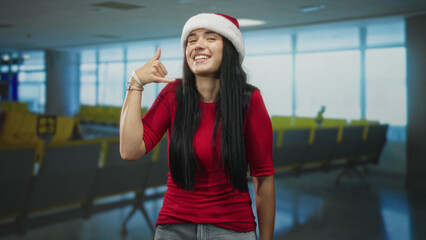 Fototapeta premium Young woman displays call me hand gesture with pointing finger in airport seating area wearing santa hat and red shirt, smiling broadly; holiday joy.