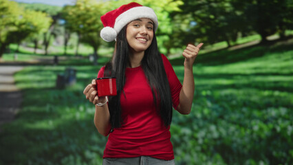 Fototapeta premium Young woman wearing santa hat holding red mug and giving thumbs up in forest; holiday cheer cheerful.