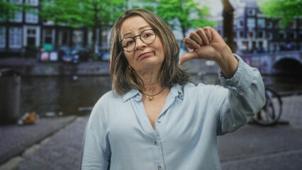 Fototapeta premium Hispanic woman with gray hair and glasses gives thumb down gesture on a street by a canal, wearing light blue shirt and necklace; disapproval reflection.