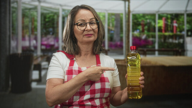 Hispanic woman cook points finger at bottle of cooking oil while holding it in a building kitchen wearing a red checkered apron; home cooking confidence.