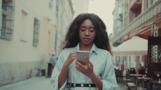 Beautiful young Afro-American woman in trendy outfit walks in the city passing by local cafes, texts via her phone, browsing the internet. Using online map, gps. Tourism. Female portrait