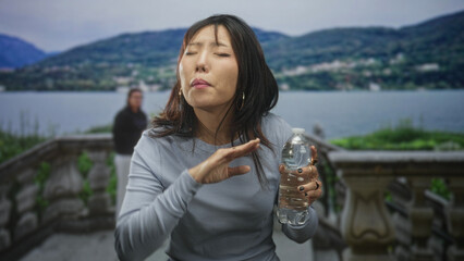 Obraz premium Young woman holding water bottle, hand at throat, coughing on a building terrace by a lake; discomfort recovery.