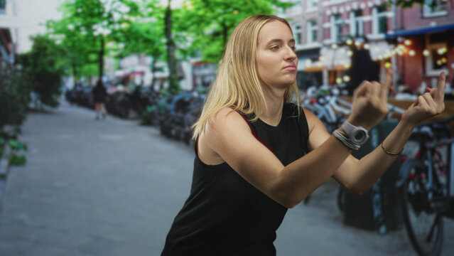 Woman showing middle finger with both hands on a city street lined with bicycles and cafe facades; defiance rebellion.