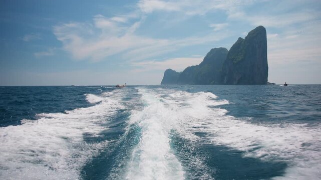 Speedboat wake creates white foam trail in turquoise Andaman Sea near Ao Pi Le in Phi Phi Thailand. Dramatic limestone karst cliffs rise from ocean under blue sky with wispy clouds on sunny day.