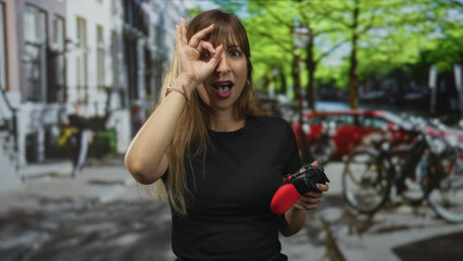 Woman holding red gamepad and making ok sign to eye on city street with parked bicycles and cars in view  curiosity. © Krakenimages.com
