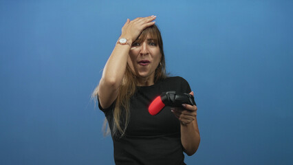 Woman gripping black and red game controller with hand on forehead and long hair visible in studio  surprise reaction. © Krakenimages.com