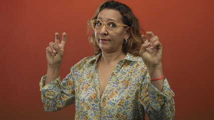 Obraz premium Middle aged hispanic woman making air quotes with fingers in studio portrait against orange backdrop; skepticism irony.
