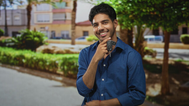 Man holds car keys by bare hand near chin on street while smiling and relaxed; confidence casual ease.
