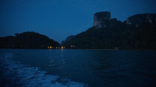 Boat wake churns through dark ocean waters at dusk as towering limestone karst cliffs covered in dense tropical jungle rise dramatically against deep blue twilight sky with distant shore lights.