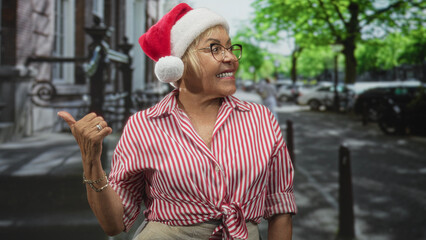 Fototapeta premium Woman wearing santa hat and glasses points thumb over shoulder on a leafy city street while smiling in a striped tied shirt and beige trousers; holiday joy.
