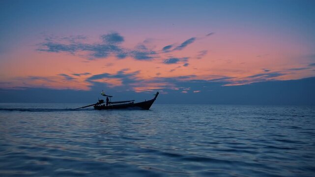 Slow motion footage of traditional Thai longtail boat silhouette cruising across calm rippling ocean waters during stunning pink and blue sunset twilight hour with scattered clouds in Mueang Krabi.