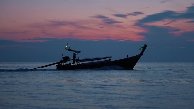 Silhouette of traditional Thai longtail boat with glowing navigation lights gliding across calm ocean waters at dusk with dramatic pink and purple clouds reflecting on the serene blue sea surface.