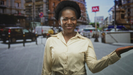Fototapeta premium Woman presenting palm with open hand gesture on a busy city street near a metro entrance, smiling at camera and gesturing; welcoming guidance.