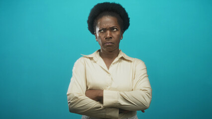 Fototapeta premium Woman with crossed arms and a frown standing centered in a studio portrait against a turquoise backdrop; disapproval resolve authority.