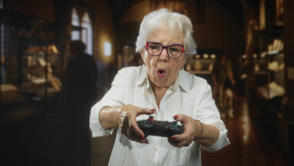 Senior woman holding a gamepad with both hands, thumbs on buttons, focused expression in a museum exhibit hall  playful engagement. © Krakenimages.com