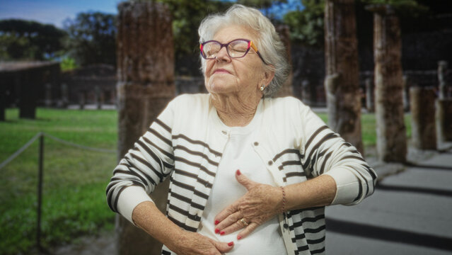 Senior woman clutching chest and abdomen, eyes closed, wearing striped cardigan and glasses, standing among ancient columns at a building site; pain concern vulnerability.