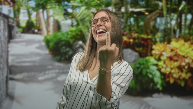 Woman wearing glasses and striped shirt smiles and displays middle finger gesture in forest setting; defiance rebellion.
