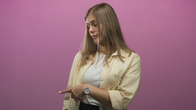 Woman with blonde hair and unimpressed expression wearing yellow shirt and white top points finger to side in pink studio; skepticism questioning.