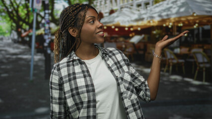 Fototapeta premium Woman with braided hair wearing white tee and plaid shirt points index finger up on restaurant terrace under string lights; confidence reminder.