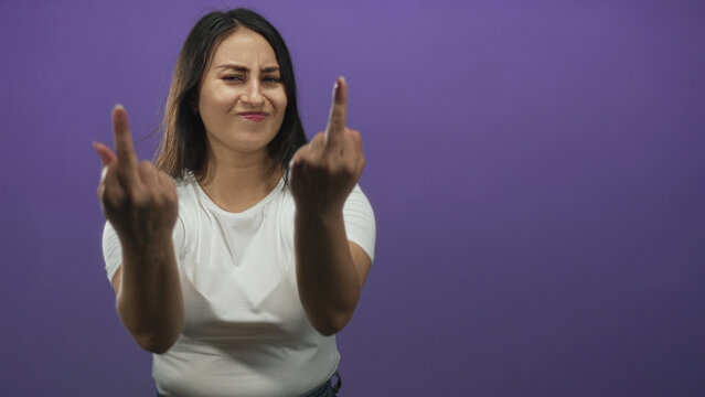 Young hispanic woman shows both middle fingers with bare hands in purple studio wearing white t shirt and jeans; defiance attitude.