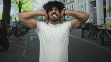 Fototapeta premium Man covering ears with hands on a city street, grimacing with muscular arms visible against loud traffic noise; urban noise stress.