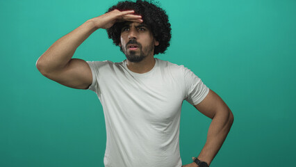 Fototapeta premium Man with curly hair shading forehead with hand in turquoise studio wearing white t shirt; curiosity.