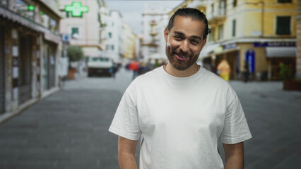Fototapeta premium Man smiling in a white t shirt, standing on a busy city street with blurred shops and pedestrians; casual warmth.
