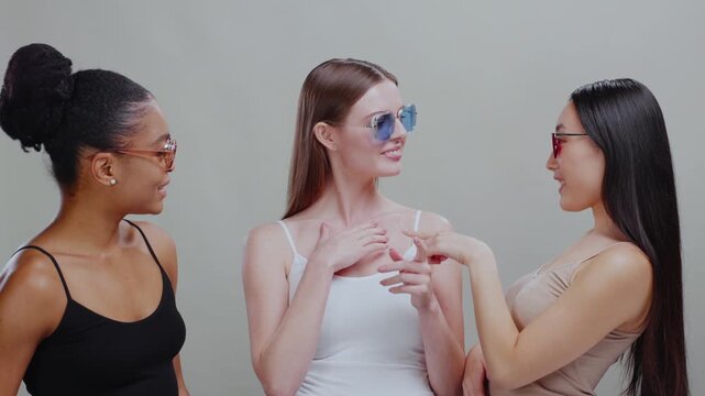 Group of amazing stylish skincare models multi-ethnic young women trying different sunglasses smiling having fun at grey background. Accessories.