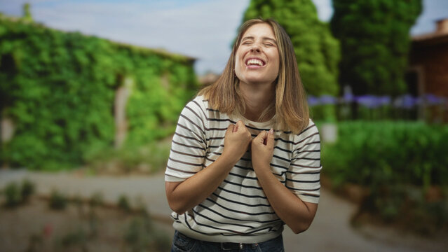 Young woman pumps fists with bare forearms visible, wide smile, standing on a garden path by a building; celebration joy.