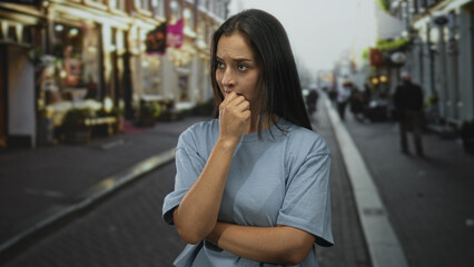 Fototapeta premium Young hispanic woman biting fingernails on a city street, hand to mouth and arms crossed; anxiety uncertainty.