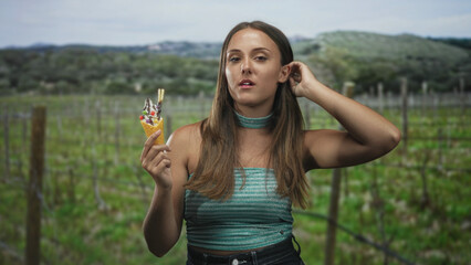 Naklejka premium Woman holding ice cream cone with hand to mouth gesture, bare forearm visible, wearing sleeveless top and choker, looking at camera while standing in a forest vineyard row; disgust.