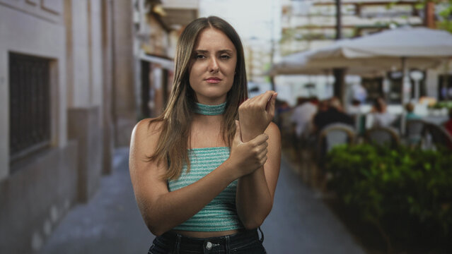 Young brunette woman holding and rubbing her wrist with a pained expression, wincing while standing near an outdoor cafe on a street; pain injury.