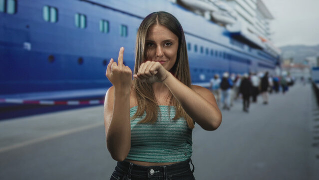 Young woman shows middle finger and clenches fist at port beside a blue cruise ship, bare arms visible; defiance attitude.