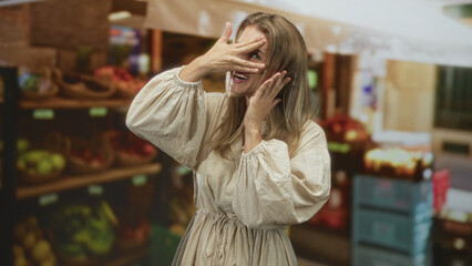 Fototapeta premium Blonde woman covers her face with a hand while smiling at a market stall in a building; playful surprise.