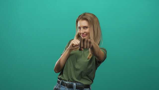 Woman with fists raised in playful boxing pose, smiling with mock punch, wearing green tshirt and jeans in studio greenscreen backdrop; playful confidence.