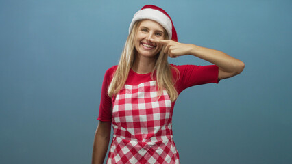 Obraz premium Woman wearing santa hat and red checked apron points finger to nose while smiling in a studio with plain blue backdrop; playful holiday cheer.