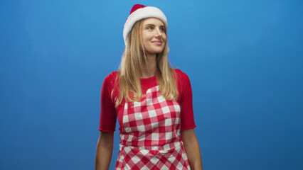 Obraz premium Woman wearing santa hat and red gingham apron smiles and looks right while tilting her head in a bright studio; cheerful holiday warmth.