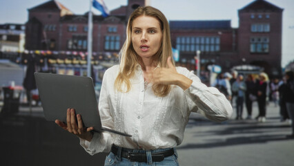 Fototapeta premium Blonde woman in white blouse and jeans holding laptop, right hand to throat speaking gesture on crowded street in front of brick building; concentration instruction presentation.