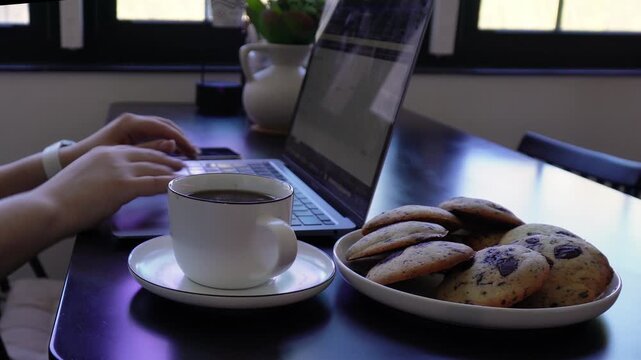 Female hands working on laptop at home with coffee cup and cookies on black desk.
