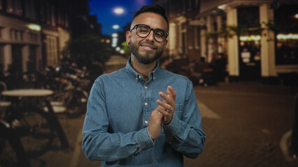 Obraz premium Man clapping hands and smiling on a bustling city street at night, wearing denim shirt, glasses and silver rings visible; celebration joy.