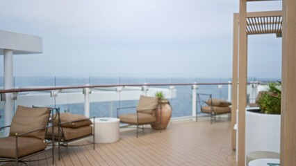 Fototapeta premium Blurred deck with lounge chairs, glass railing, planters and distant ocean horizon in soft focus outdoor; background backdrop copyspace.