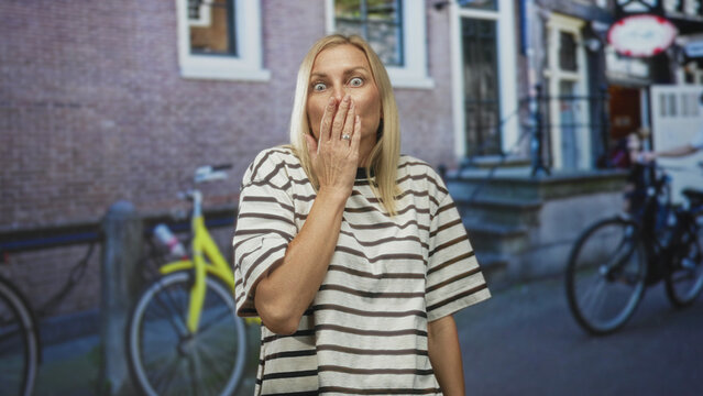 Woman with hand covering mouth showing surprised expression on a city street with bicycles and brick building; astonishment candid moment.