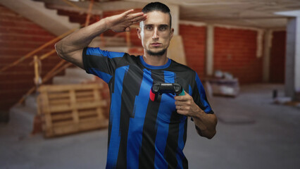 Man holding a gamepad with both hands and saluting while wearing a blue soccer jersey in an unfinished building with staircase and wooden pallets  playful. © Krakenimages.com