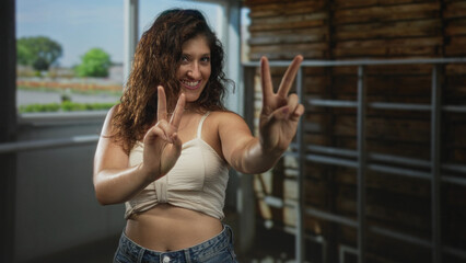Obraz premium Woman showing double peace signs and bare midriff in studio space with window light and wooden rack; joy celebration.